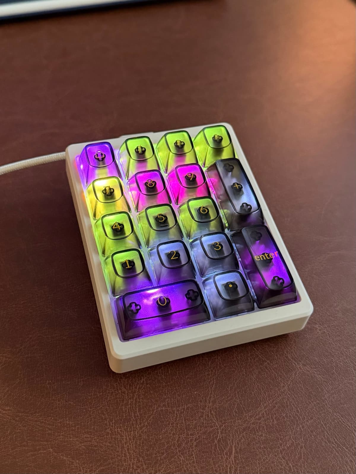 a white magicforce numpad lit up on a brown leather desk mat. violet heartbeat top left, lime keys working, a gold call shouting, a hot pink reply, the wide zero glowing violet. the printed legends still visible under the light.
